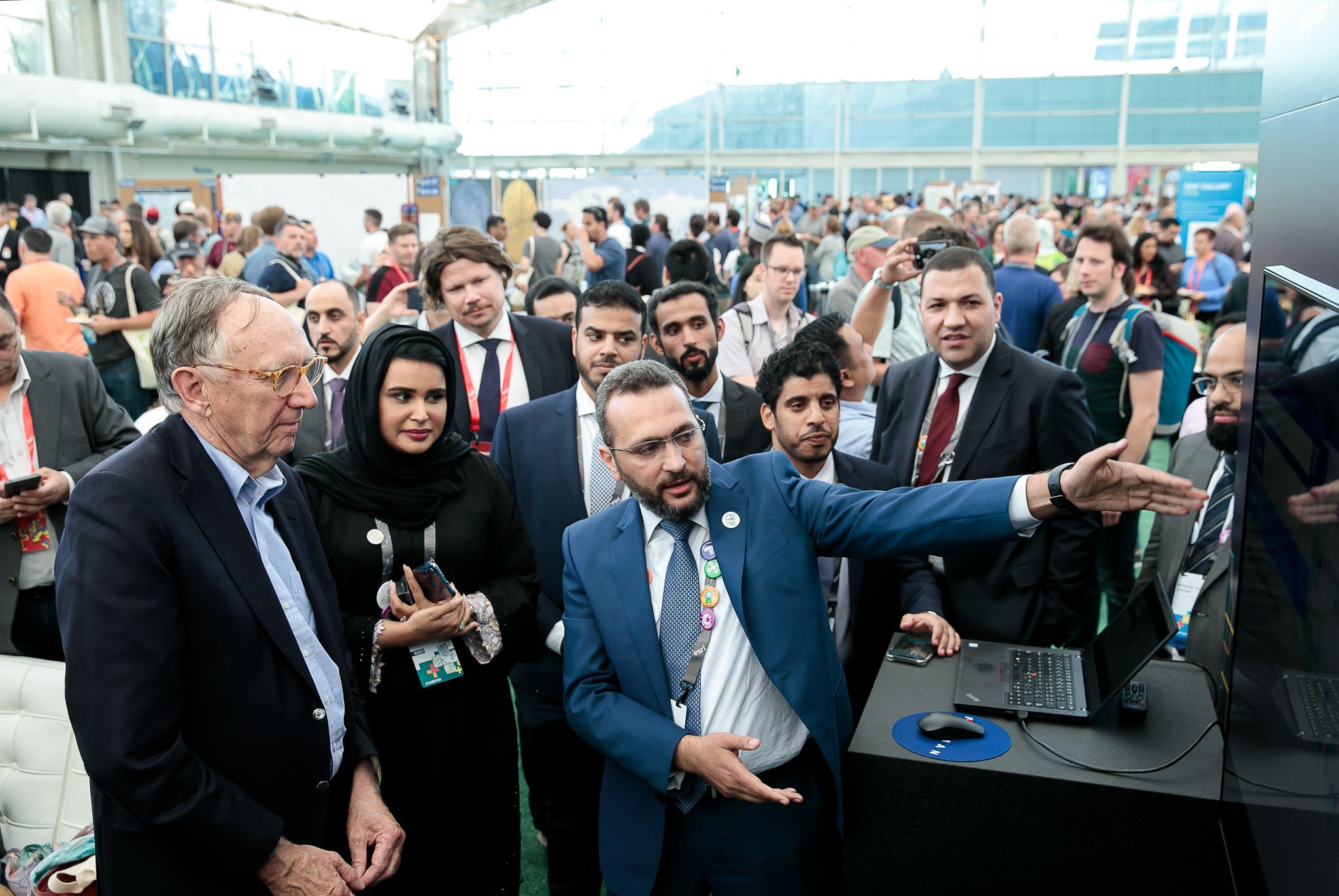 A diverse group of attendees is engaged in a presentation or demonstration led by a man in a blue suit and glasses