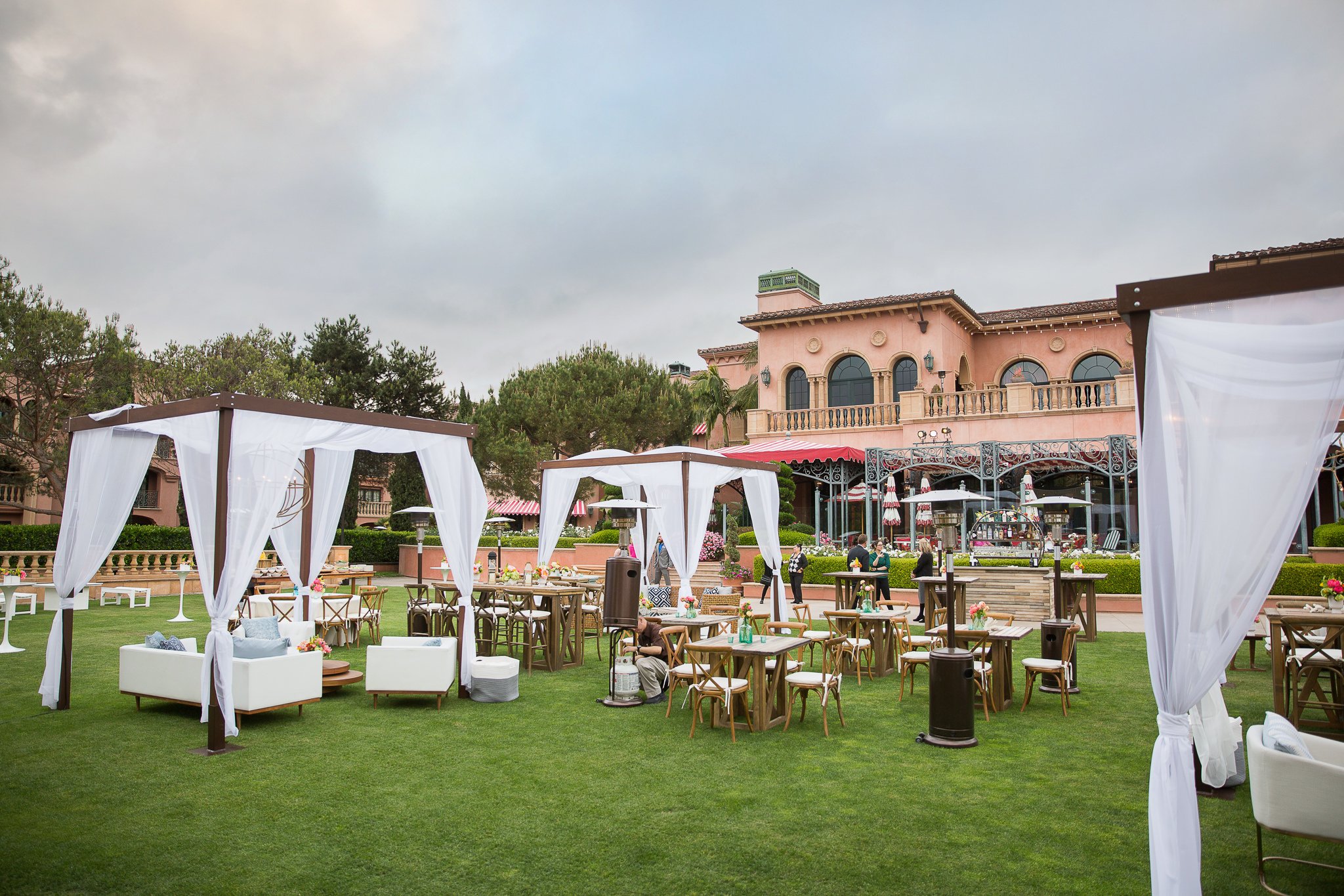 An elegant outdoor event setup on a well-manicured lawn in front of a luxurious Mediterranean-style building with arched windows, ornate balconies, and red-tiled roofs