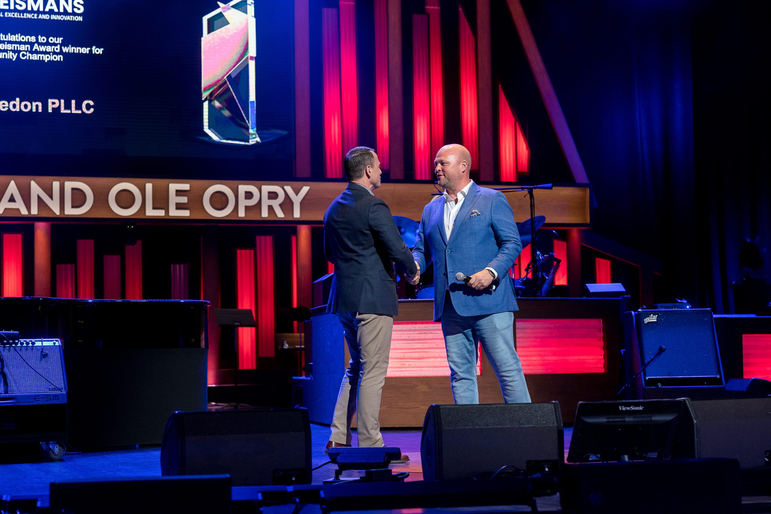 Two men are shaking hands, one wearing a dark suit and the other in a blue blazer with jeans, holding a microphone