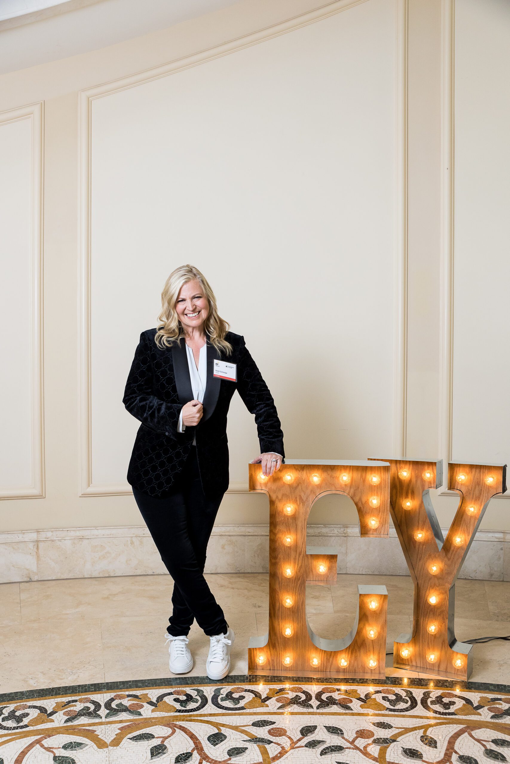 A woman in a velvet blazer posing next to light-up "EY" letters at an event.