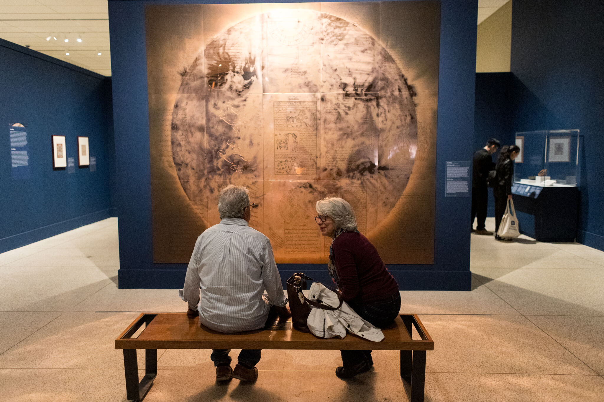 Two people sit on a bench, admiring a large painting displayed in front of them