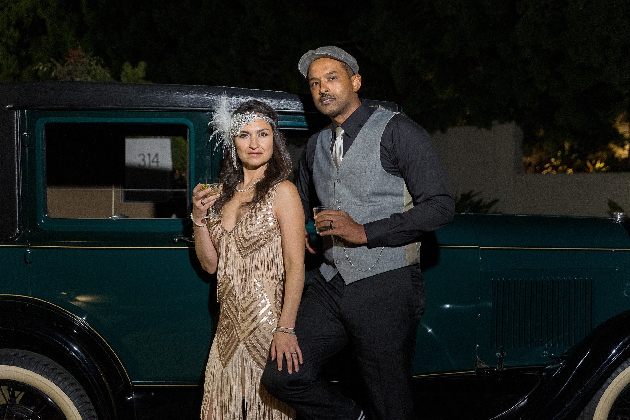A man and woman in 1920s attire pose beside a vintage car