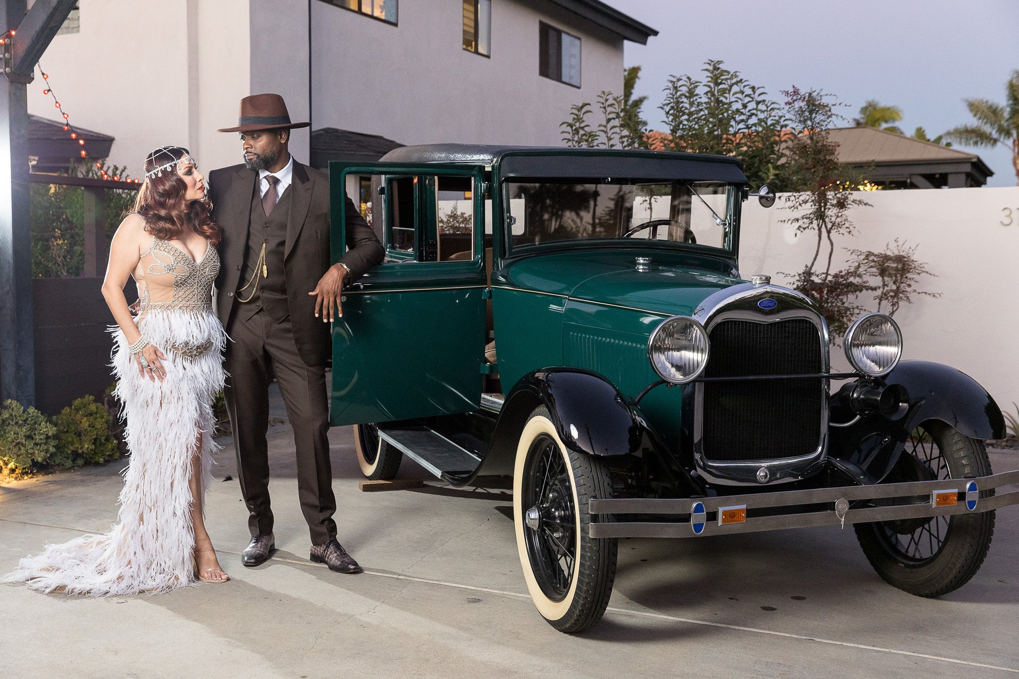 A couple smiles while posing with a vintage car in front of a charming house