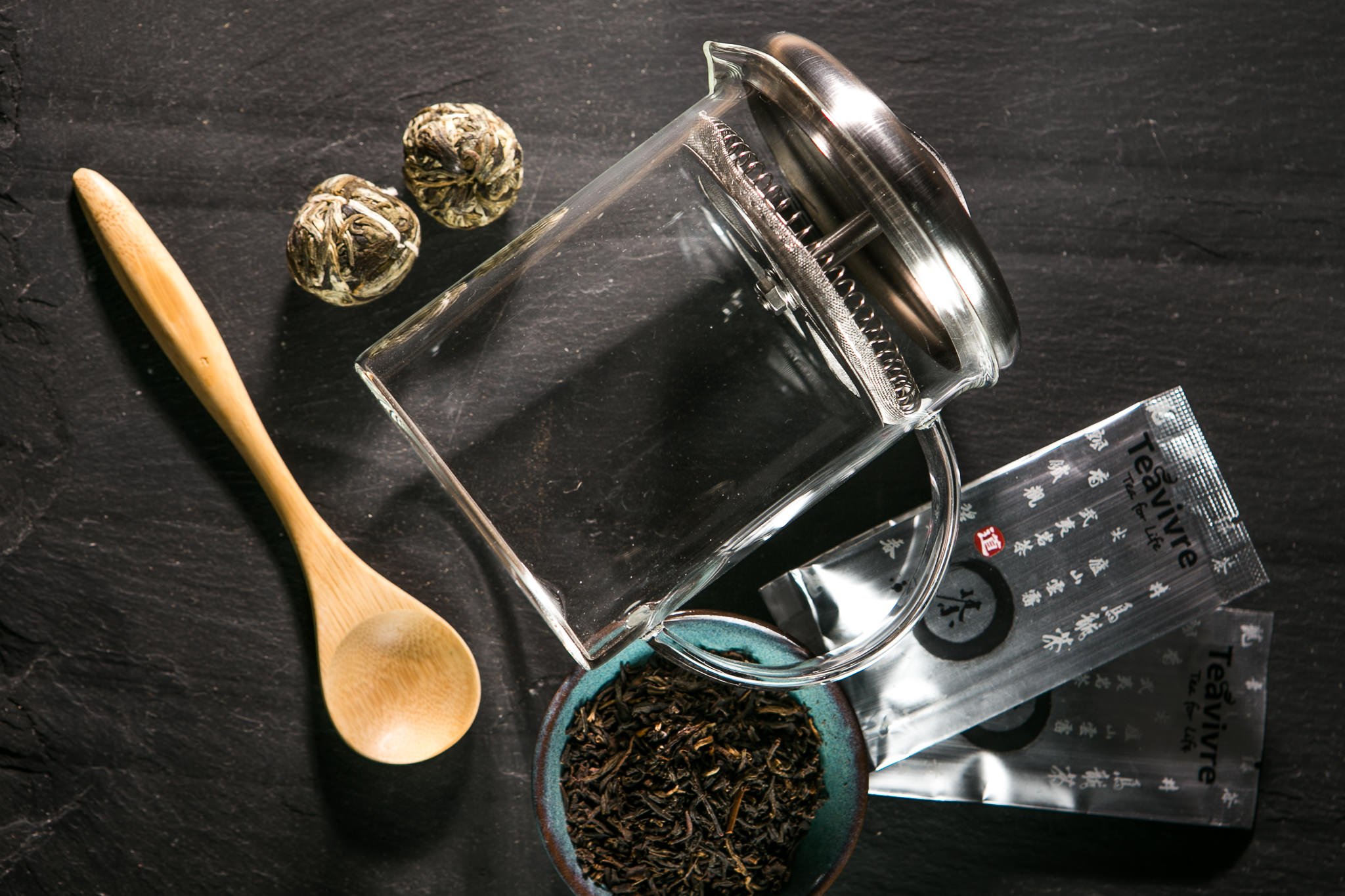 A flat lay of a tea setup, including a glass tea infuser, loose leaf tea, a wooden spoon, and tea packets