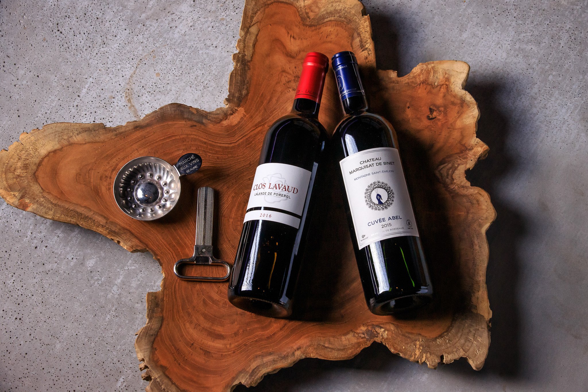 A flat lay of two wine bottles, a wine opener, and a wine pourer on a rustic wooden slab