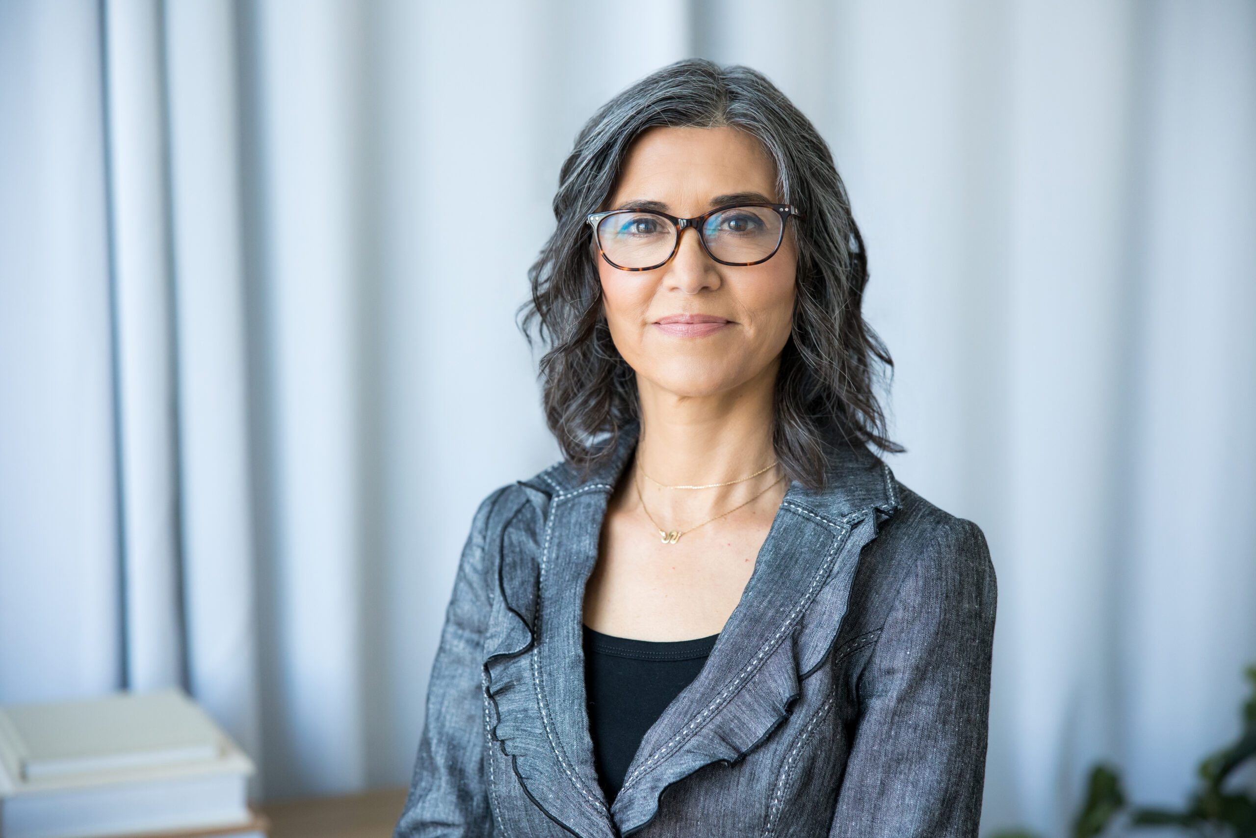 A woman wearing glasses and a gray jacket, looking confidently at the camera