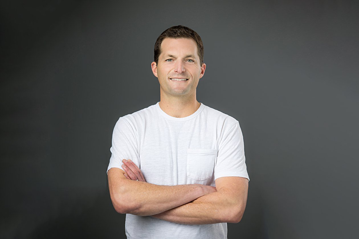 A man in a white t-shirt stands with his arms crossed, displaying a confident posture