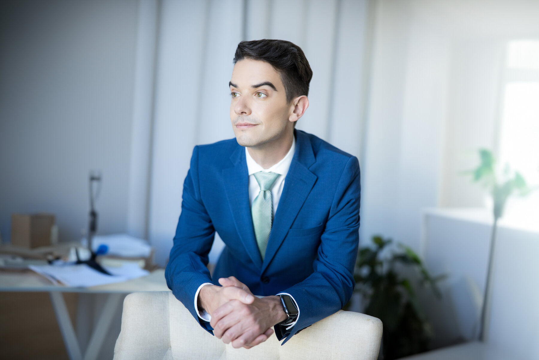 A young man in a blue suit, looking thoughtful, rests his arms on a chair in an office setting