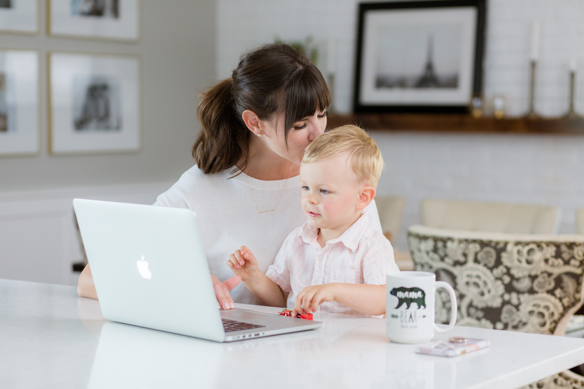 Mother working on a laptop while holding her toddler in a bright kitchen