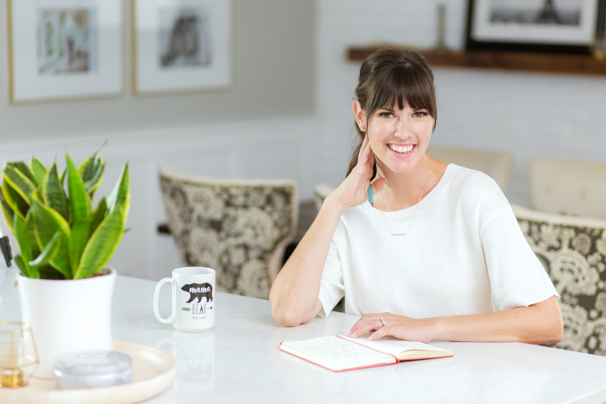 Smiling woman seated at a kitchen table with an open notebook and coffee mug