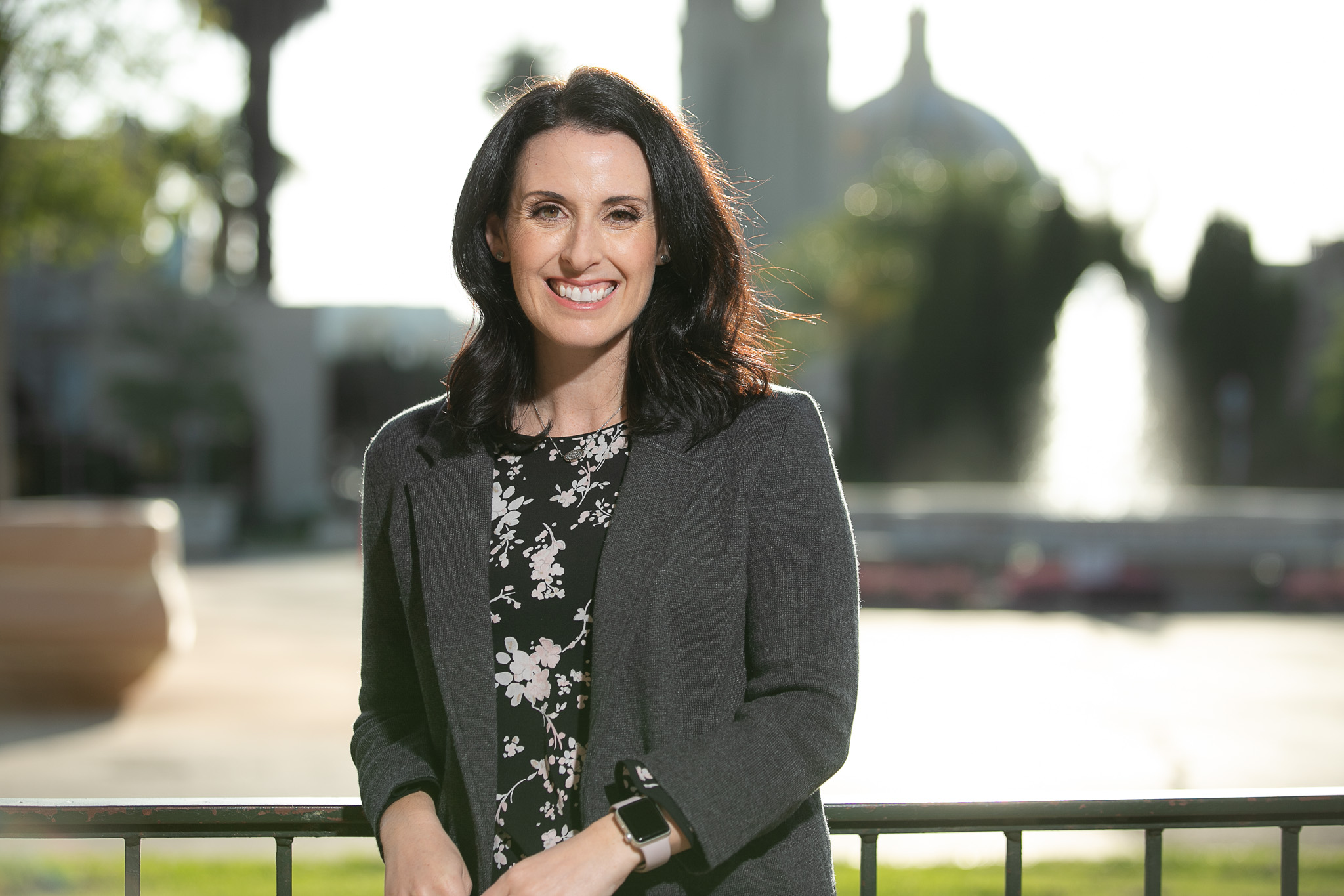 Smiling woman outdoors leaning on a railing with a blurred fountain behind her