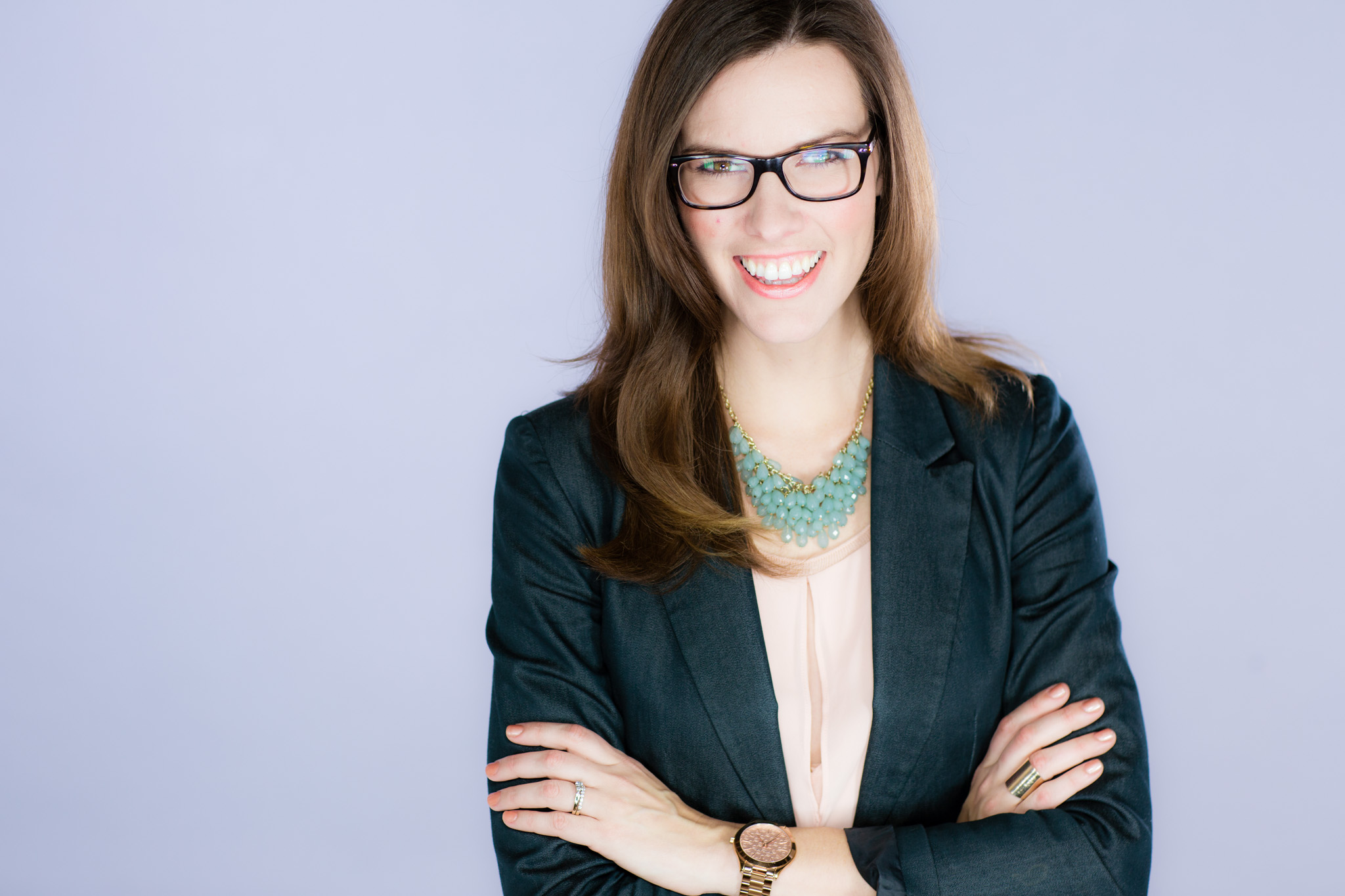 Smiling woman in glasses and blazer posing with arms crossed against a studio backdrop